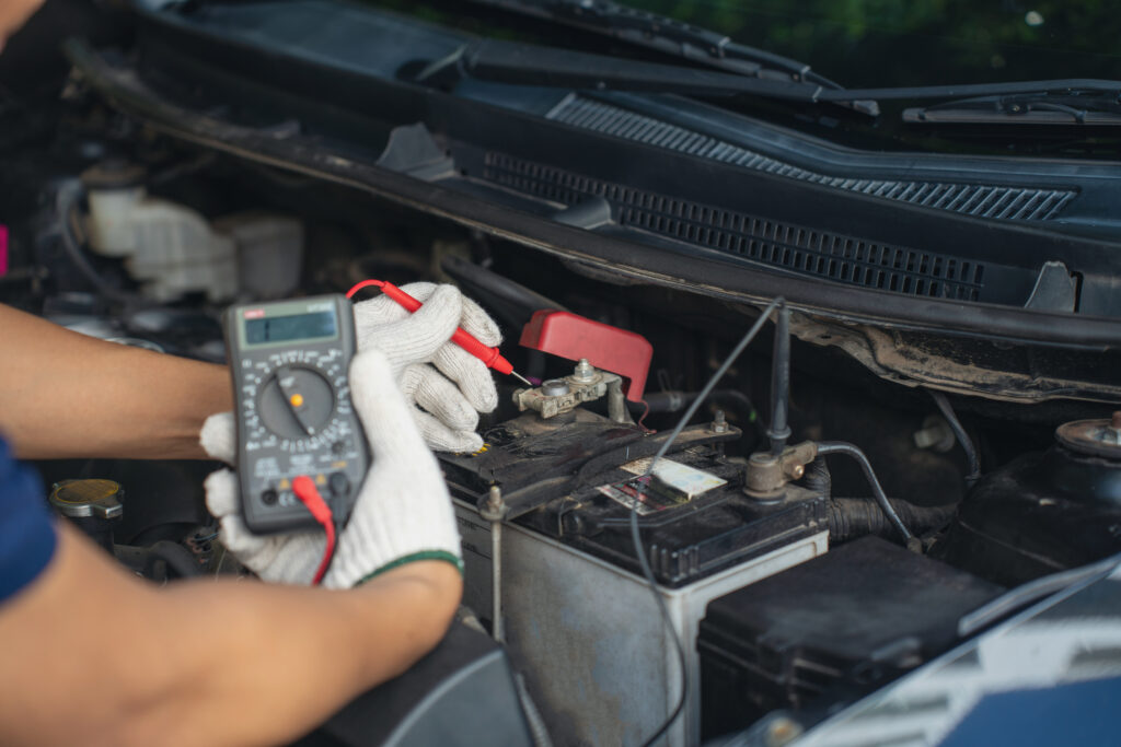 Close-up of mechanic holding voltmeter to check voltage car battery energy problem for service maintenance. Car mechanic noting repair parts during open car hood engine repair unrecognisable